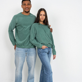  Eine Frau und ein Mann stehen beide lächelnd an einer Wand und tragen dabei den grünen Unisex Sweater von Oktopulli