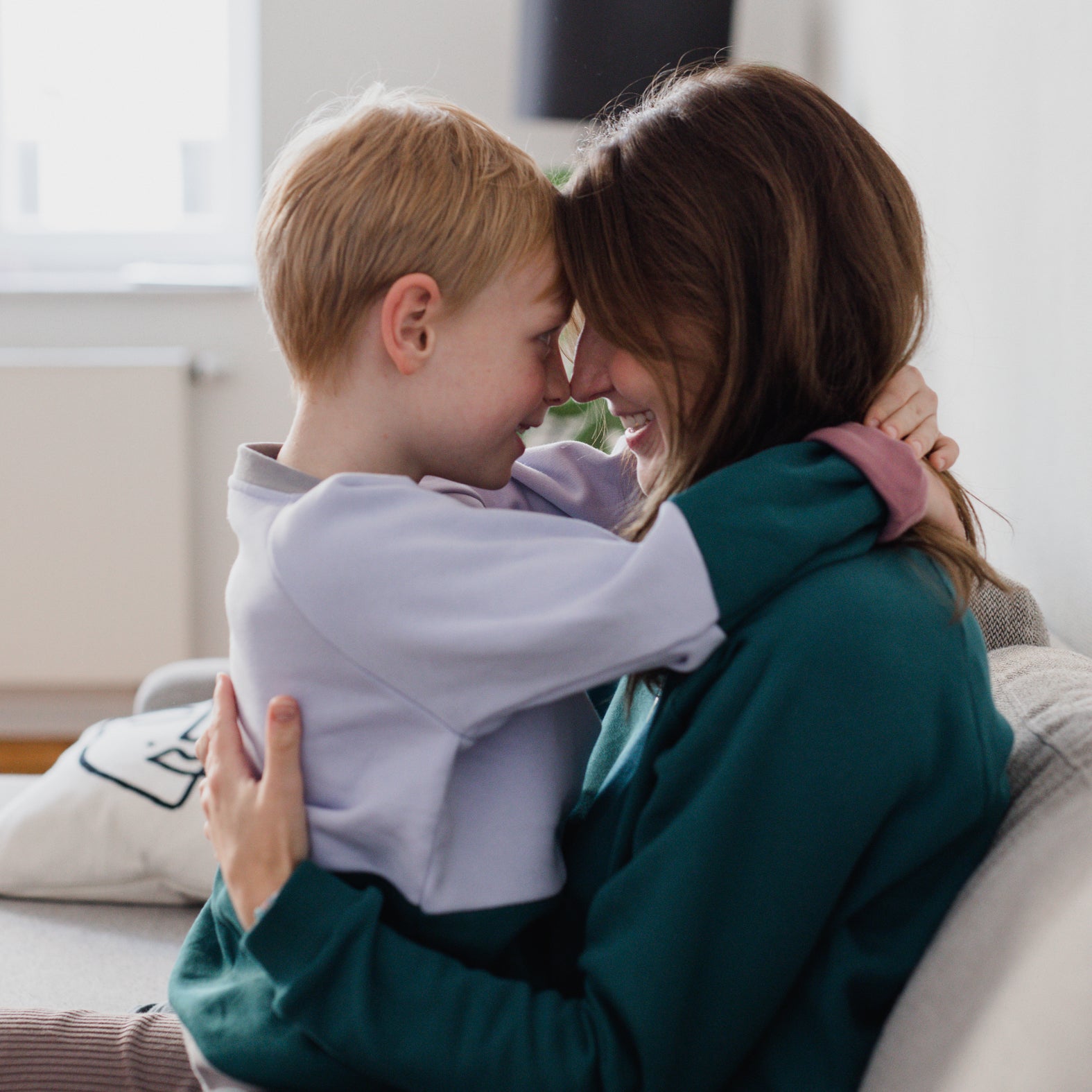 Ein Kind im grünen und fliederfarbenen Oktopulli sitzt auf dem Schoß seiner Mutter, die einen smaragdgrünen Oktopulli trägt. 